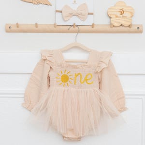 May include: A beige baby romper with long sleeves and a tulle skirt, embroidered with a yellow sun and the word "one." A matching bow headband and wooden decor are also visible.