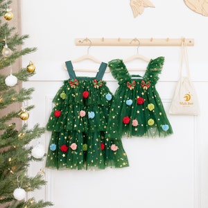 Può includere: Due abiti di albero di Natale in tulle verde con glitter dorati e pon pon colorati appesi a un rack di legno. Gli abiti hanno un fiocco sul davanti e sono a strati di tulle. Una piccola borsa bianca con il testo "Mali Boo" è appesa al rack.