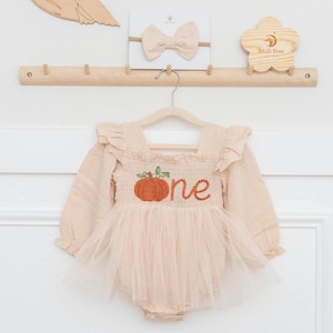 May include: A beige long-sleeve romper with ruffled shoulders and a tulle skirt overlay. The bodice features an embroidered orange pumpkin and the word one in orange cursive script. A matching beige bow headband is displayed on a wooden rack.