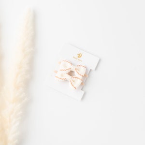 May include: Two white fabric hair bows with orange stitching on a white card with the text "Mali Boo" in gold lettering.