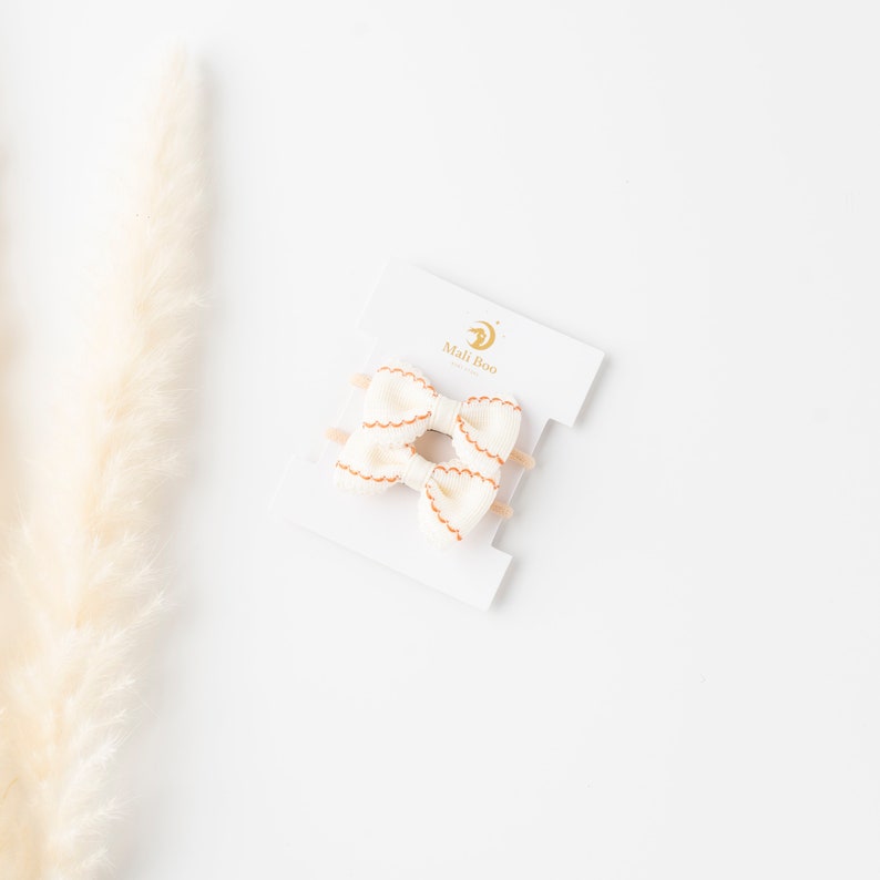 May include: Two white fabric hair bows with orange stitching on a white card with the text "Mali Boo" in gold lettering.