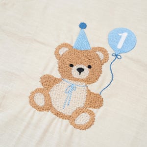May include: A beige fabric featuring an embroidered brown teddy bear wearing a blue party hat. The bear has a cream-colored belly and holds a blue balloon with a white number 1 stitched in the center, connected by a thin blue thread.