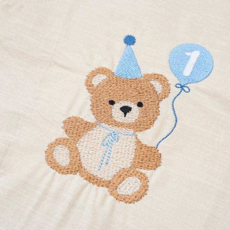 May include: A beige fabric featuring an embroidered brown teddy bear wearing a blue party hat. The bear has a cream-colored belly and holds a blue balloon with a white number 1 stitched in the center, connected by a thin blue thread.