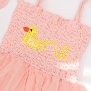 May include: A light pink baby dress with a yellow embroidered duck and the word "one". The dress has a smocked bodice, spaghetti straps, and a tulle skirt. The duck has an orange beak.