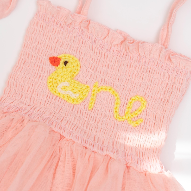 May include: A light pink baby dress with a yellow embroidered duck and the word "one". The dress has a smocked bodice, spaghetti straps, and a tulle skirt. The duck has an orange beak.