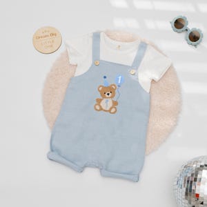 May include: A light blue overall romper with a white short-sleeve t-shirt underneath. The romper features an embroidered brown teddy bear wearing a blue party hat and holding a blue balloon with the number 1. Two wooden buttons secure the straps.