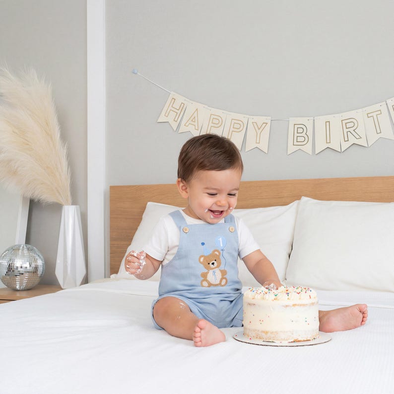 May include: A light blue overall short set with a white t-shirt underneath. The overalls feature an embroidered brown teddy bear holding a blue balloon with the number one printed on it. The outfit has wooden buttons at the shoulder straps.