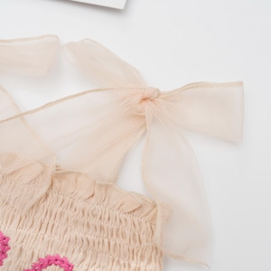 May include: Close-up of a beige top with pink floral embroidery. The top features smocking and delicate, sheer, peach-colored ribbon straps tied into bows at the shoulders. The fabric has a soft, textured appearance.