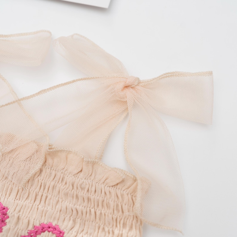 May include: Close-up of a beige top with pink floral embroidery. The top features smocking and delicate, sheer, peach-colored ribbon straps tied into bows at the shoulders. The fabric has a soft, textured appearance.