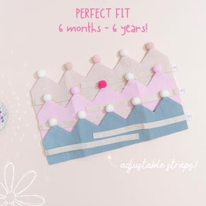 May include: A stack of fabric crowns in shades of pink and blue, each with a pom-pom on top. The crowns have adjustable straps and are designed for children aged 6 months to 6 years. The text "PERFECT FIT" is visible.