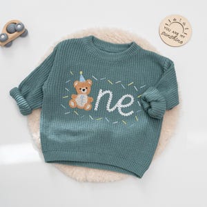 May include: Teal knit sweater with a teddy bear and the word "one" embroidered in white. The teddy bear wears a party hat. The sweater is on a white, fluffy surface. A wooden toy car and a wooden sign that says "YOU ARE MY sunshine" are in the background.