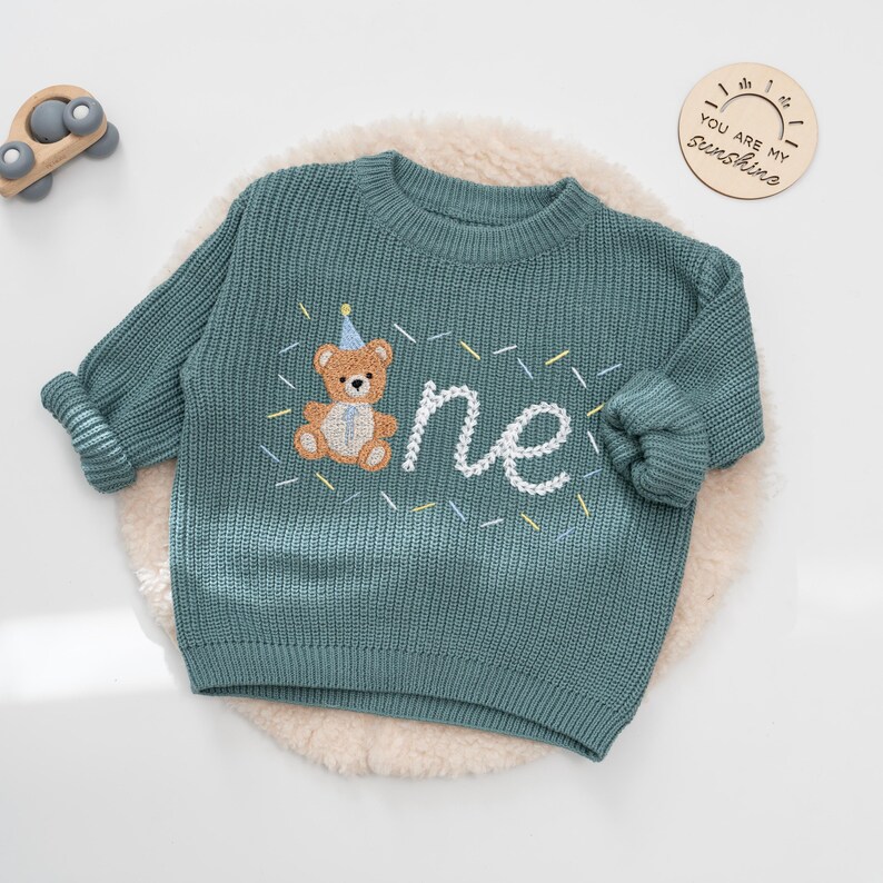May include: Teal knit sweater with a teddy bear and the word "one" embroidered in white. The teddy bear wears a party hat. The sweater is on a white, fluffy surface. A wooden toy car and a wooden sign that says "YOU ARE MY sunshine" are in the background.