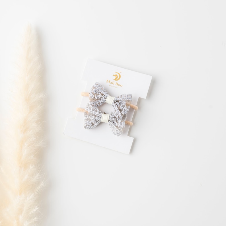 May include: Two silver glitter hair bows on a white card with the text "Mali Boo" in gold lettering.