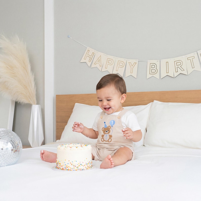May include: A beige romper featuring an embroidered brown teddy bear wearing a blue party hat and holding a blue balloon with the number one. The romper is worn with a white short-sleeved shirt. A white cake with colorful sprinkles sits nearby.