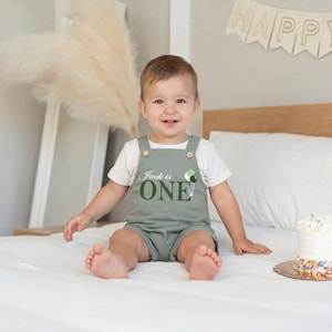 May include: A child wearing a sage green romper with the text "Jack is ONE" and a balloon graphic. The child is sitting on a white bed, smiling. A small cake with sprinkles is on a wooden plate.