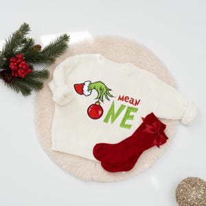May include: Cream-colored knit sweater with a Grinch-themed design, featuring a green hand holding a red ornament and the words "Mean One" in green. Paired with red socks with a bow. Festive holiday apparel.