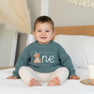 May include: A teal knitted sweater with the word "one" embroidered in white, featuring a teddy bear with a party hat. The baby is wearing cream-colored pants and is sitting on a white bed.