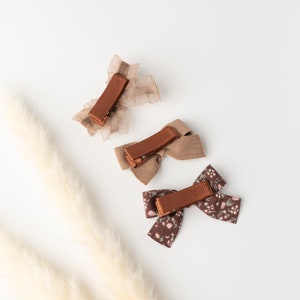 May include: Three brown hair clips with different designs. One is a brown bow with a floral pattern, one is a brown bow with a plain fabric, and one is a brown ribbon with a sheer fabric.
