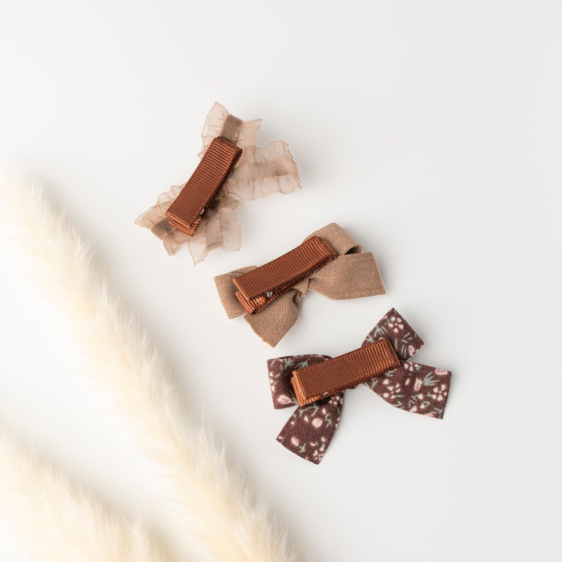 May include: Three brown hair clips with different designs. One is a brown bow with a floral pattern, one is a brown bow with a plain fabric, and one is a brown ribbon with a sheer fabric.