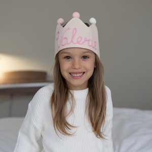 May include: A light pink fabric crown with the name "Valerie" embroidered in pink script. The crown has five points and is adorned with three small pom-poms in pink and white. The crown is worn by a smiling child.