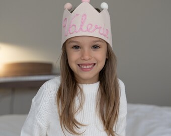 May include: A light pink fabric crown with the name "Valerie" embroidered in pink script. The crown has five points and is adorned with three small pom-poms in pink and white. The crown is worn by a smiling child.