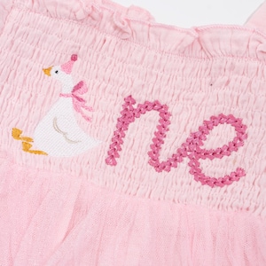 May include: Pink baby dress with a ruffled neckline and a tulle skirt. The dress features an embroidered goose wearing a party hat and bow, with the word "one" embroidered in pink thread.
