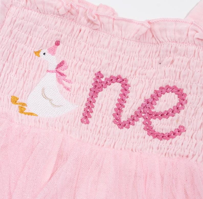 May include: Pink baby dress with a ruffled neckline and a tulle skirt. The dress features an embroidered goose wearing a party hat and bow, with the word "one" embroidered in pink thread.