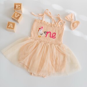 May include: A peach-colored baby romper with a tulle skirt and a matching bow. The romper features a chicken applique and the word "one" embroidered on the bodice. Wooden blocks spell out "EVA".