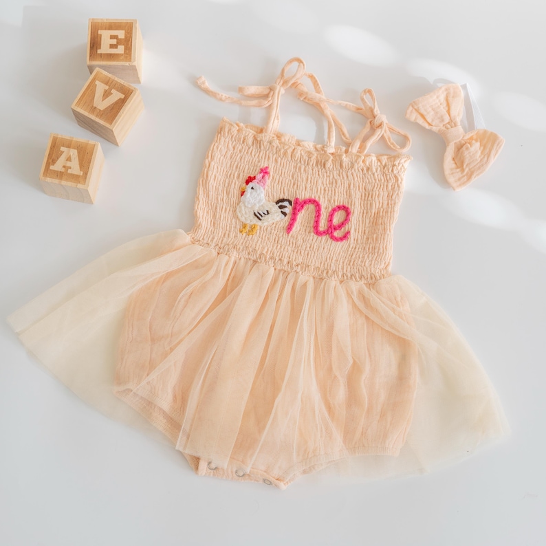 May include: A peach-colored baby romper with a tulle skirt and a matching bow. The romper features a chicken applique and the word "one" embroidered on the bodice. Wooden blocks spell out "EVA".