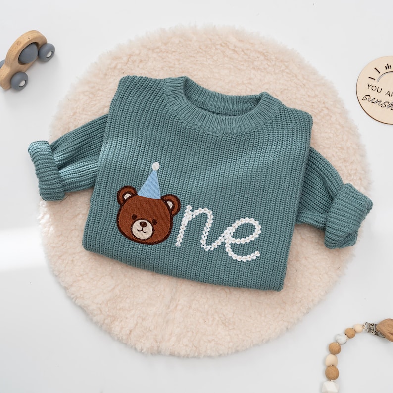 K&ouml;nnte beinhalten: Ein teal-farbener Strickpullover mit langen &Auml;rmeln. Der Pullover hat einen gestickten braunen B&auml;ren mit einer blauen Partyh&uuml;tchen und das Wort "one" in wei&szlig;er Schrift. Der Pullover liegt auf einem cremefarbenen, runden, flauschigen Teppich.