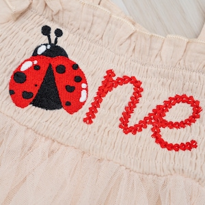 May include: Close-up of a beige garment with a red embroidered ladybug and the word "one". The ladybug has black spots and white accents. The word "one" is stitched in red thread.