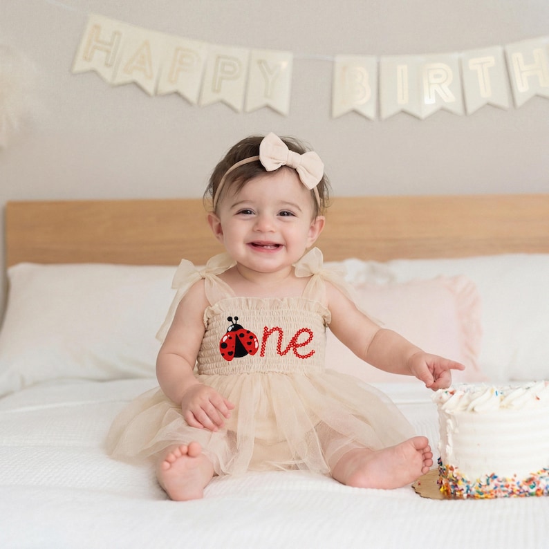 May include: A smiling baby wearing a beige ladybug-themed dress with the word "one" and a matching bow headband. A white cake with sprinkles sits nearby, and a banner that says "Happy Birthday" is in the background.