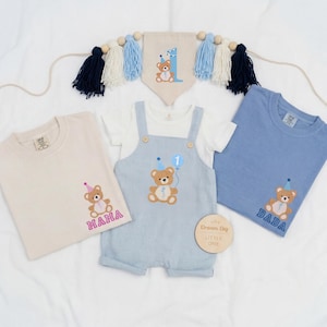 May include: A beige t-shirt with a teddy bear and the word MAMA, a light blue overall with a teddy bear and the number 1, a blue t-shirt with a teddy bear and the word DADA, and a pennant banner with a teddy bear and the number 1 with tassels.