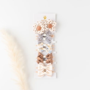 May include: Set of 7 hair clips for babies and toddlers. The set includes two white and brown floral clips, two blue and white floral clips, two brown satin bows, and one white and brown scalloped bow. The clips are displayed on a white card with the text "Mali Bo Baby" printed on it.