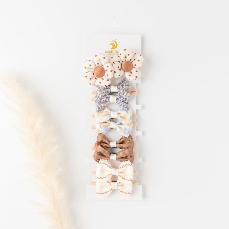 May include: Set of 7 hair clips for babies and toddlers. The set includes two white and brown floral clips, two blue and white floral clips, two brown satin bows, and one white and brown scalloped bow. The clips are displayed on a white card with the text "Mali Bo Baby" printed on it.