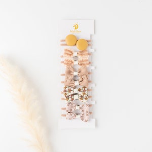 May include: A set of eight hair bows in various shades of brown and beige, displayed on a white card with the text "Mali Boo Baby Shop". The bows are made of different fabrics, including floral print, textured fabric, and solid colors. Two yellow hair clips are also included.