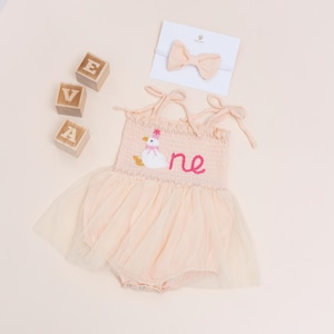 May include: A peach-colored baby romper with a tulle skirt, embroidered with a duck wearing a pink party hat and the word "one" in pink. A matching bow headband and wooden blocks spelling "EVA" are also included.