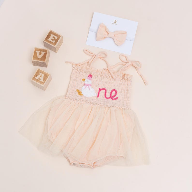 May include: A peach-colored baby romper with a tulle skirt, embroidered with a duck wearing a pink party hat and the word "one" in pink. A matching bow headband and wooden blocks spelling "EVA" are also included.