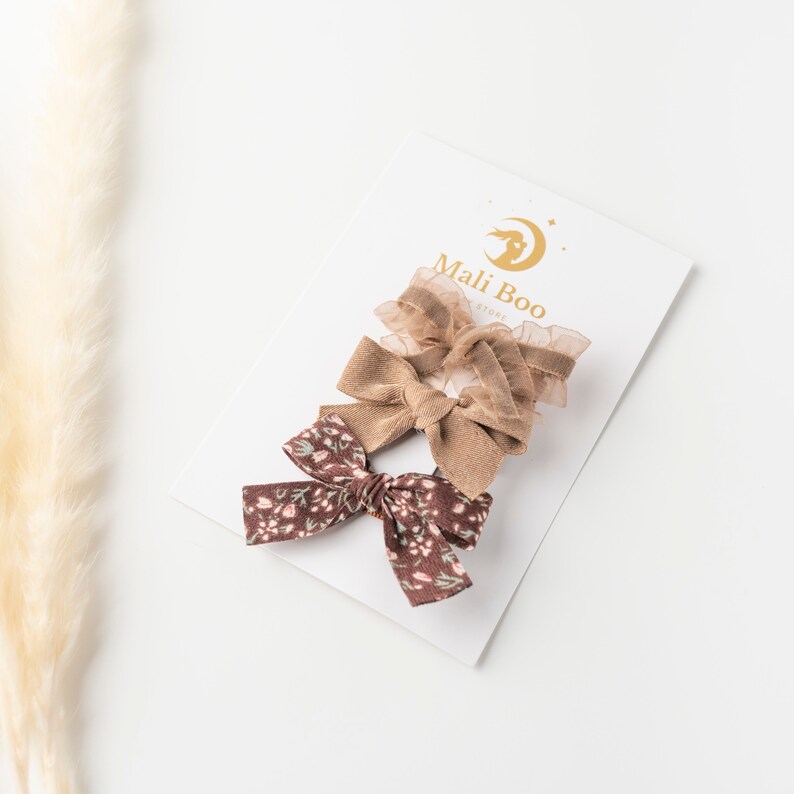 May include: Three hair bows on a white card with the text "Mali Boo Store". The bows are brown and floral patterned.