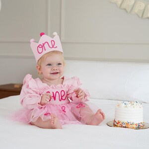 May include: A baby wearing a pink dress and a pink crown with the word "one" embroidered on it. The baby is sitting on a white bed with a small cake nearby. The cake has white frosting and colorful sprinkles.