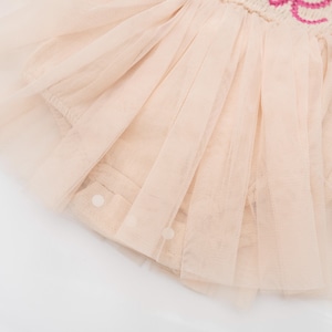 May include: Close-up of a light pink tulle skirt with a layered design. The skirt has a soft, flowing appearance, with multiple layers of sheer fabric. The garment is adorned with small, round buttons and a decorative embroidered detail.