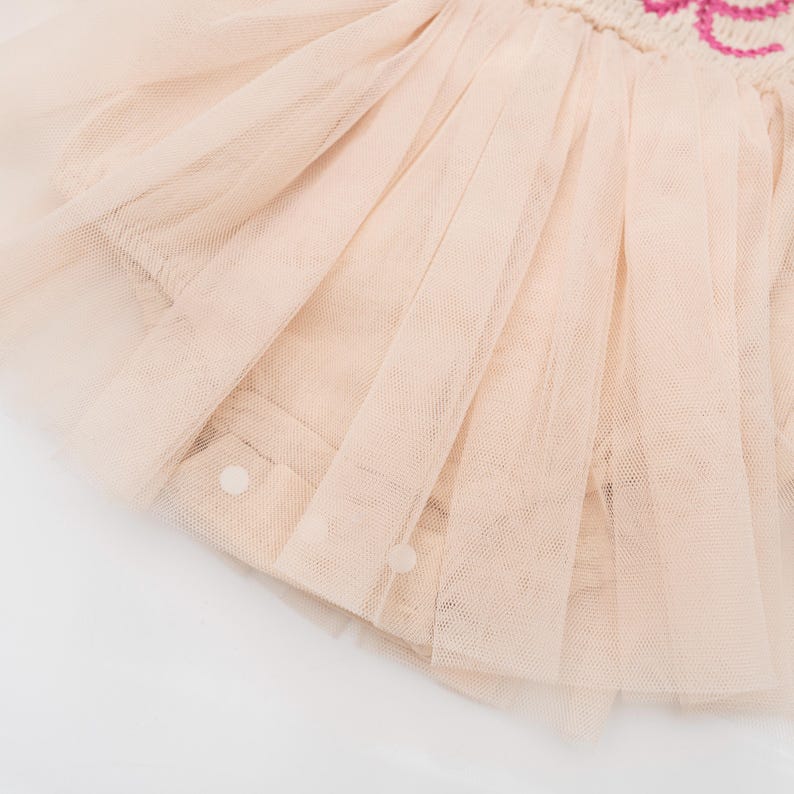 May include: Close-up of a light pink tulle skirt with a layered design. The skirt has a soft, flowing appearance, with multiple layers of sheer fabric. The garment is adorned with small, round buttons and a decorative embroidered detail.