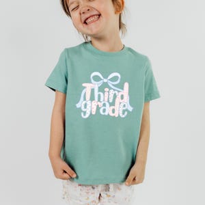 May include: A mint green t-shirt with the words "Third Grade" in a playful font, accented with a bow and ribbons. The text features a gingham pattern and pastel colors. The shirt is short-sleeved.