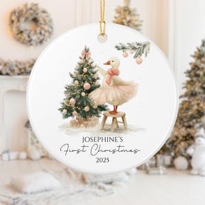 May include: A round white Christmas ornament with a gold hanging loop. It depicts a duck in a tutu decorating a Christmas tree. The text reads "Josephine's First Christmas 2025". The ornament is a festive decoration.