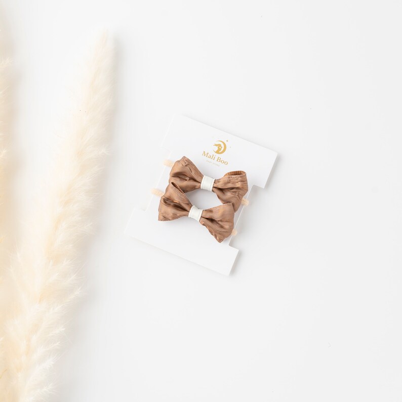 May include: Two brown satin hair bows with white centers on a white card with the text "Mali Boo" in black.