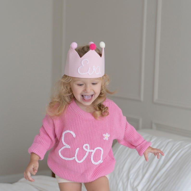 May include: A pink crown with the name "Eva" embroidered on the front, adorned with three pom-poms. The child is wearing a pink sweater with the name "Eva" and a flower embroidered on it.