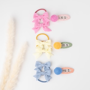 May include: Three sets of hair accessories on a white background. Each set includes a bow on a hair tie and a matching snap clip. Colors include pink, yellow, and blue bows with coordinating clips in pink, green, and orange.