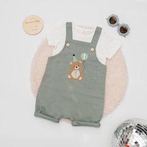 May include: A sage green overall romper with a white short-sleeve t-shirt. The romper features an embroidered brown teddy bear wearing a green party hat and holding a green balloon with the number one. Two wooden buttons secure the straps.
