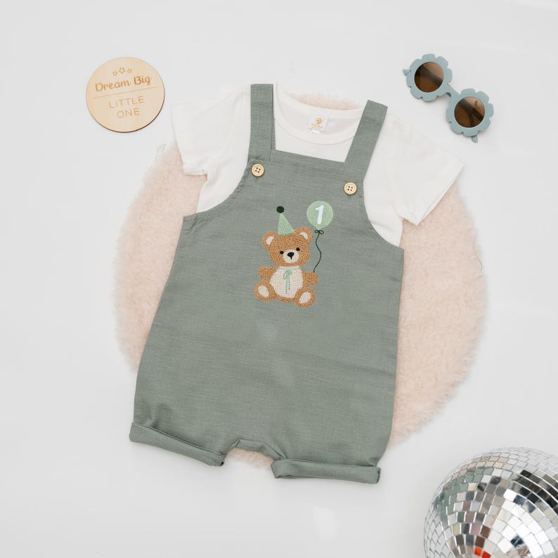 May include: A sage green overall romper with a white short-sleeve t-shirt. The romper features an embroidered brown teddy bear wearing a green party hat and holding a green balloon with the number one. Two wooden buttons secure the straps.