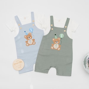 May include: Two short-sleeved rompers with overalls, one in light blue and one in sage green, each layered over a white t-shirt. Both feature an embroidered brown teddy bear wearing a party hat and holding a balloon with the number 1.
